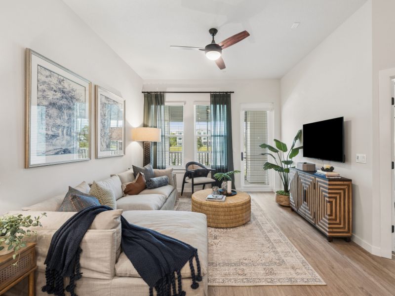 Modern living room with beige sofa, TV, round coffee table, plants, art on walls, and large windows with sheer curtains.