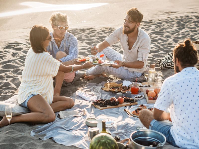 Four people enjoy a picnic on the beach at sunset, sharing food and drinks on a blanket.