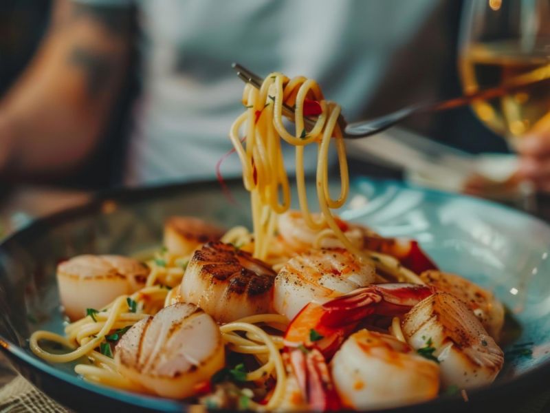 A bowl of spaghetti with seared scallops and shrimp, with a hand twirling pasta on a fork.